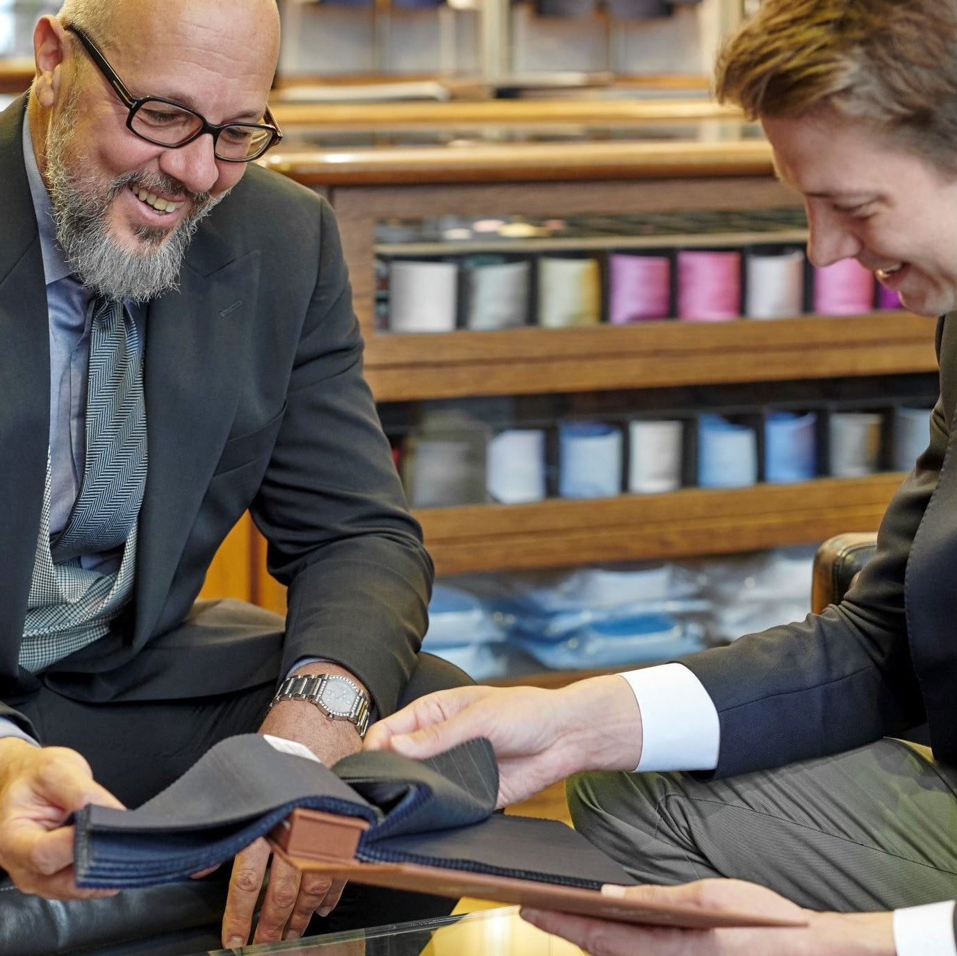 Two men in a tailor shop, selecting fabric from a large assortment, smiling and engaging in conversation.