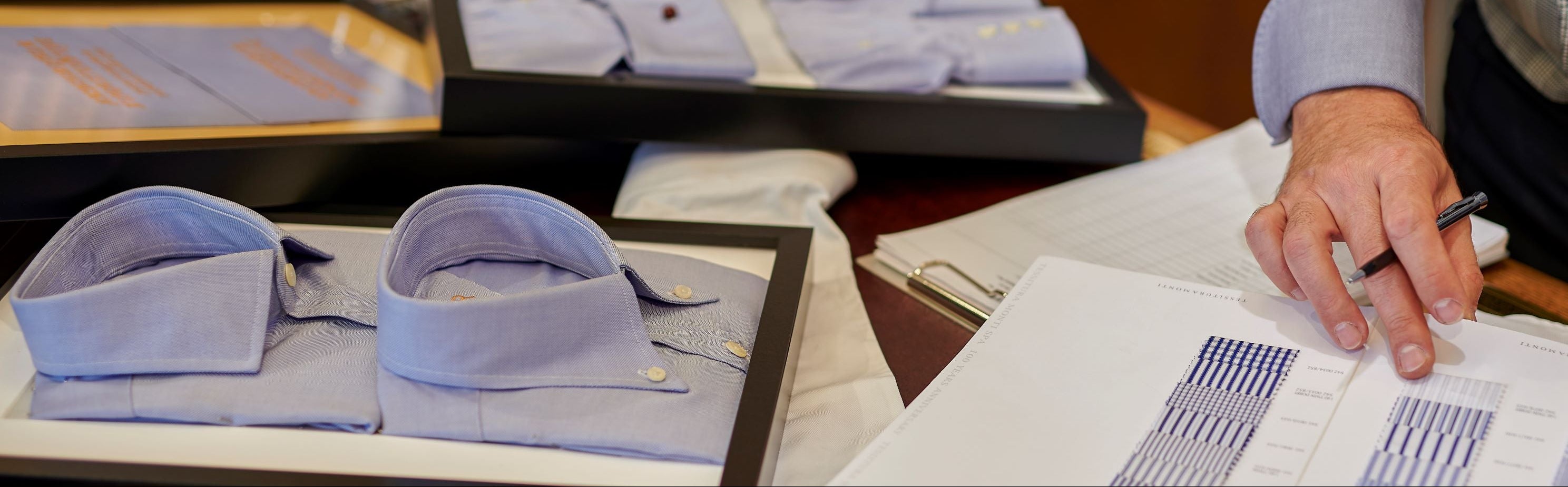 A person is measuring fabric for a bespoke shirt with a selection of ironed shirts laid out on a table.