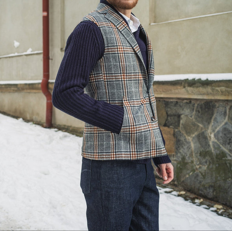 Man wearing a plaid waistcoat body warmer and sweater standing against a wall with snow on the ground.