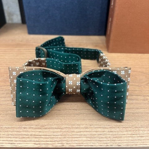 Green and brown polka dot bow tie with white accents on a wooden surface, with a blue box in the background.