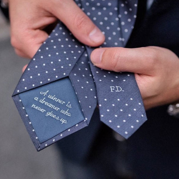 A close-up of a man holding a bespoke tie with a dotted pattern, displaying monogram initials 'P.D.' 