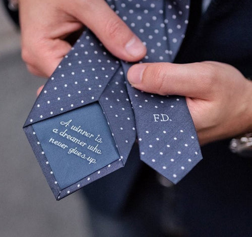 A close-up of a man holding a bespoke tie with a dotted pattern, displaying monogram initials 'P.D.' 