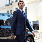 Man in a navy suit walking past a black car with a blurred building in the background
