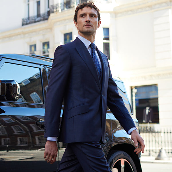 Man in a navy suit walking past a black car with a blurred building in the background